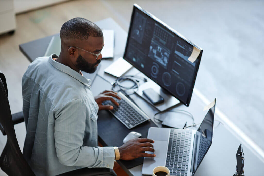 Man sitting at desk with two computers and a coffee.