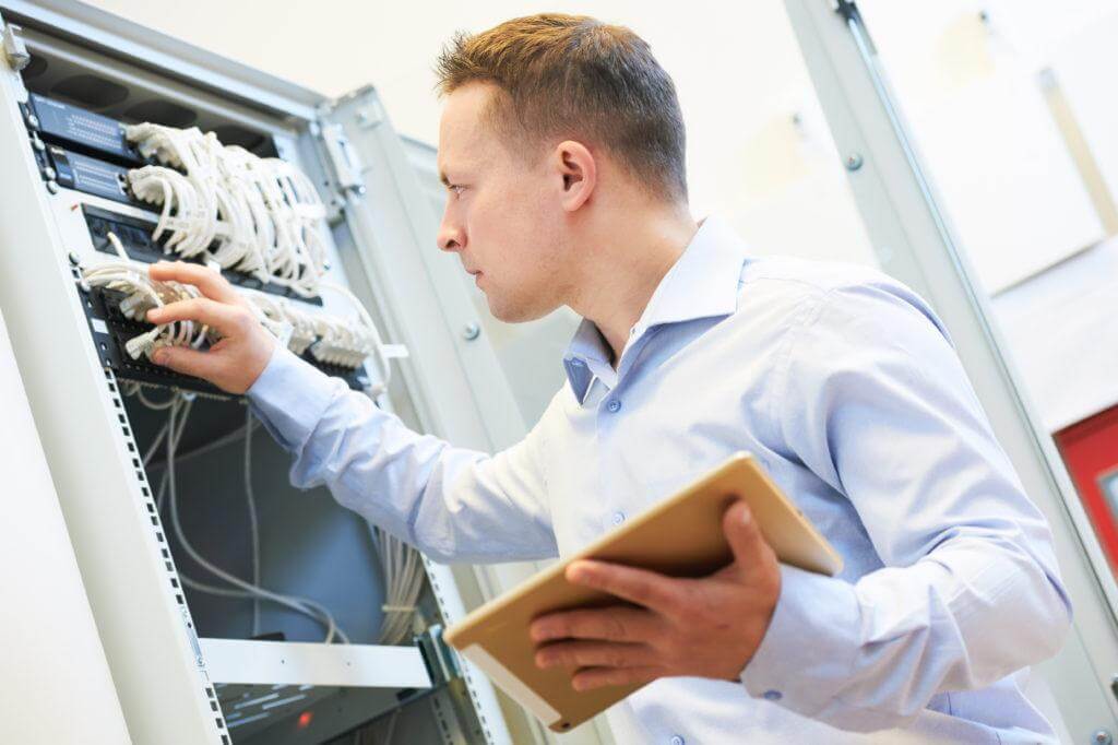 IT employee reviewing wires in a IT closet holding a tablet.