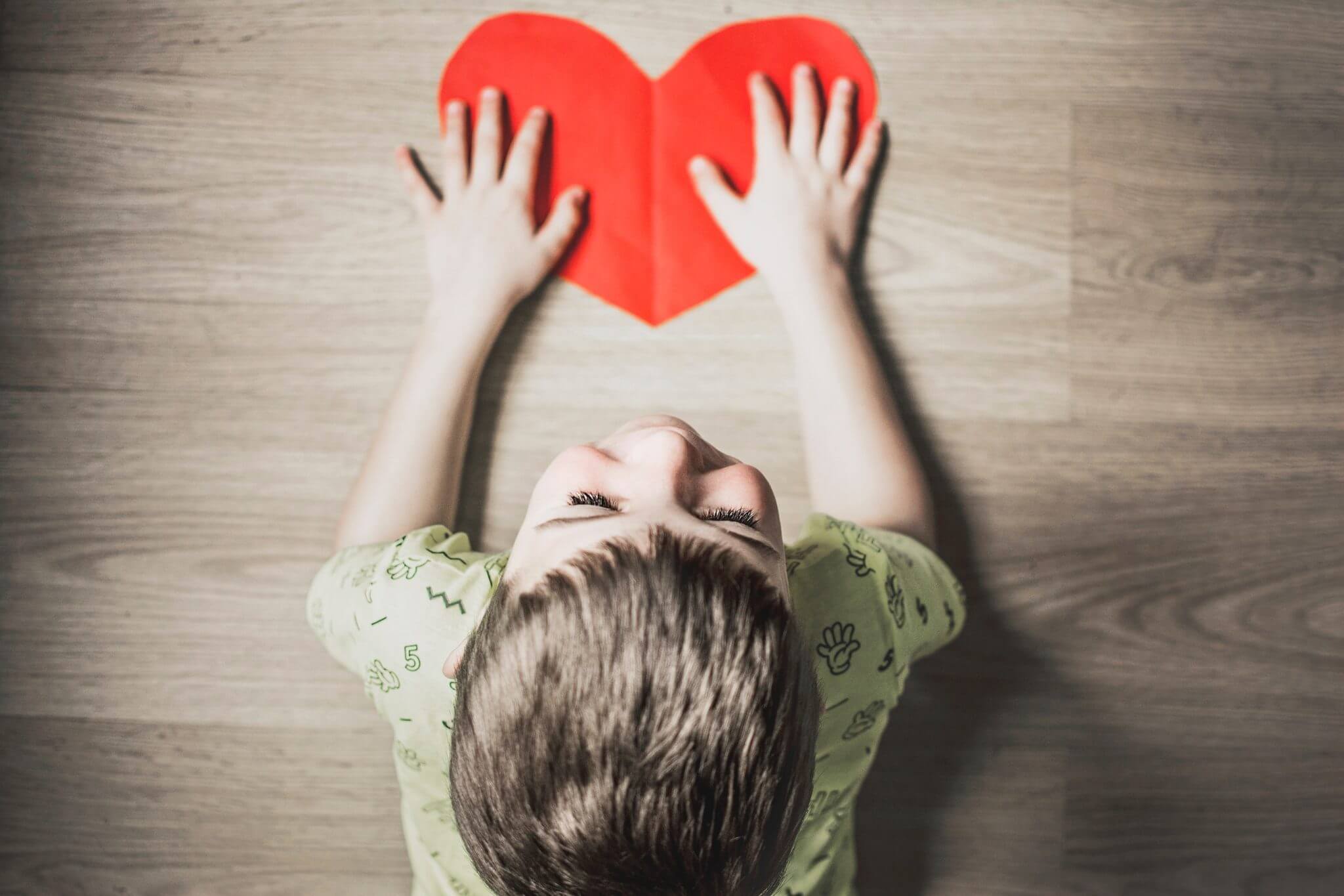 Little boy laying on the floor with hands on a red cut out paper heart.