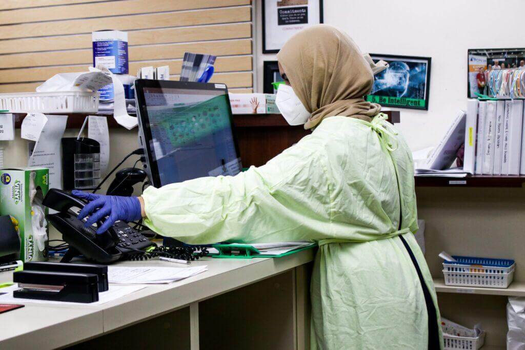 Pharmacy worker reaching for telephone while workinig on a laptop.